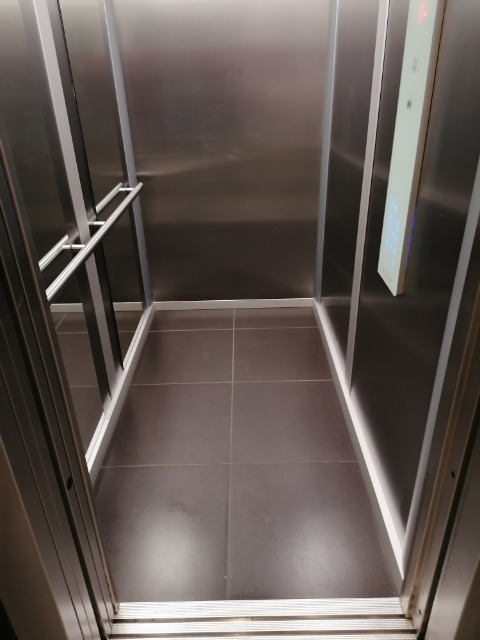 Wnętrze windy z panelami ze stali nierdzewnej, szarą podłogą z płytek i panelem sterowania z podświetlanymi przyciskami po prawej stronie.