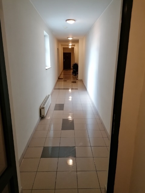 Długi korytarz wyłożony jasnymi płytkami z ciemnymi wstawkami, oświetlony punktowymi lampami sufitowymi, z grzejnikiem przy ścianie i wózkiem dziecięcym w oddali.