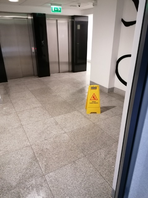 Żółty znak ostrzegawczy 'Uwaga śliska podłoga' ustawiony na szarych płytkach w biurowcu w Mińsku Mazowieckim, w tle windy i biała ściana z czarnymi dekoracjami.