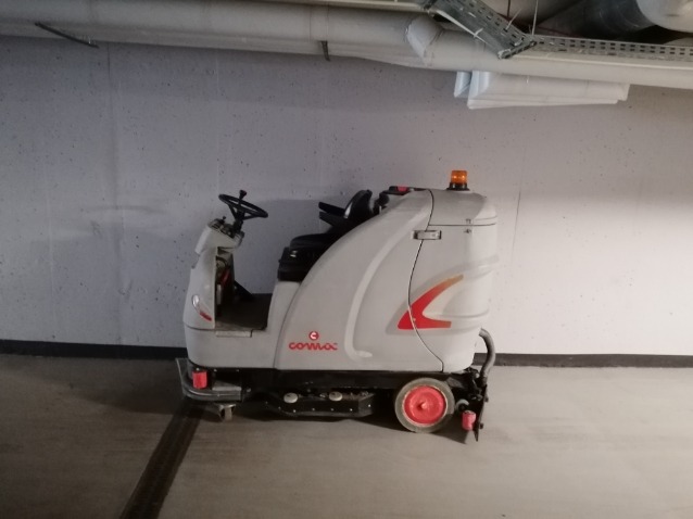 Szara samojezdna maszyna czyszcząca Comac w garażu podziemnym, widoczna pomarańczowa lampka ostrzegawcza i czerwone felgi kół.