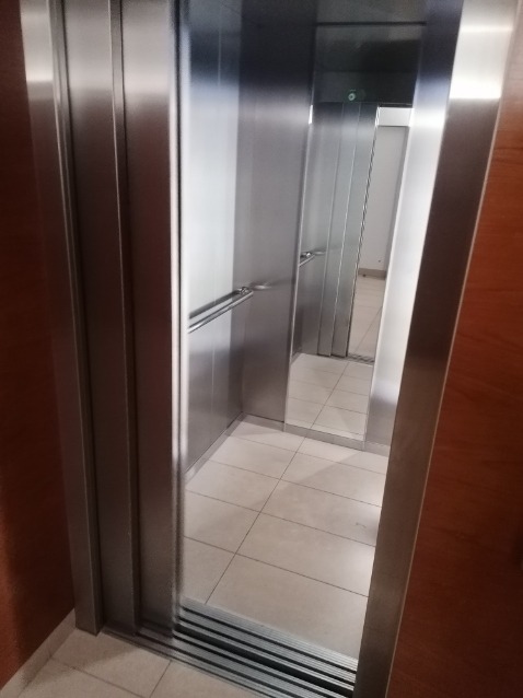 Wnętrze nowoczesnej windy osobowej z metalowymi ścianami i poręczą, widok z zewnątrz na jasną podłogę z płytek.