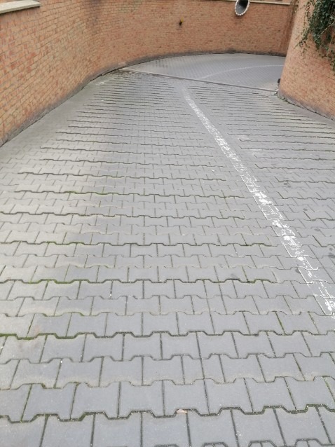 Szara kostka brukowa ułożona w łuk, z widocznymi śladami po oponach i niewielkimi chwastami między kostkami, otoczona ceglanymi ścianami.