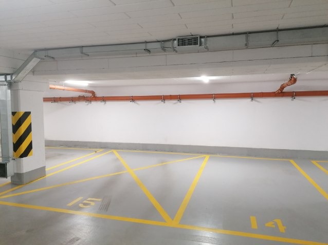 Oznakowane żółtymi liniami stanowiska postojowe w garażu podziemnym z numerami 14 i 15, widoczne rury pod sufitem i kolumna z czarno-żółtymi pasami ostrzegawczymi.