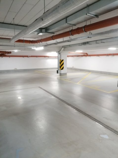 Wnętrze pustego, jasnego parkingu podziemnego z pomalowanymi żółtymi liniami na szarym betonowym podłożu, widoczne rury pod sufitem i słup z czarno-żółtymi pasami.