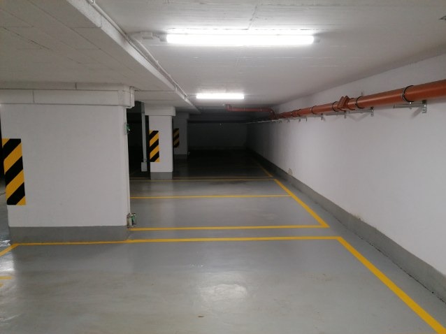 Puste, czyste, oświetlone światłem jarzeniowym podziemne miejsce parkingowe z żółtymi liniami wyznaczającymi miejsca postojowe i słupami z czarno-żółtymi pasami ostrzegawczymi.