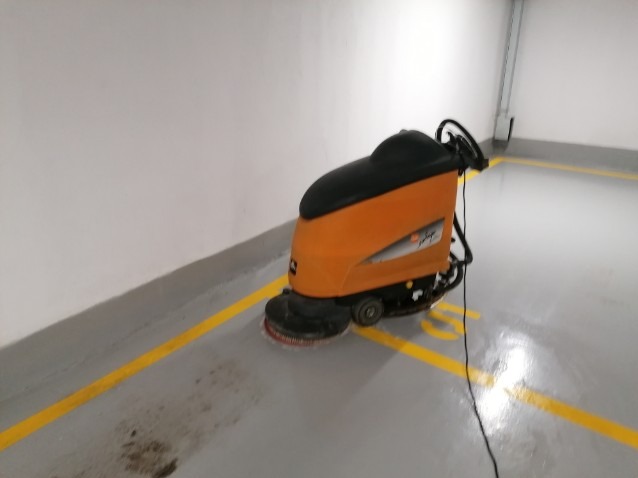 Pomarańczowa maszyna do mycia podłóg w garażu podziemnym, widoczne żółte linie na szarej posadzce i białe ściany w tle.