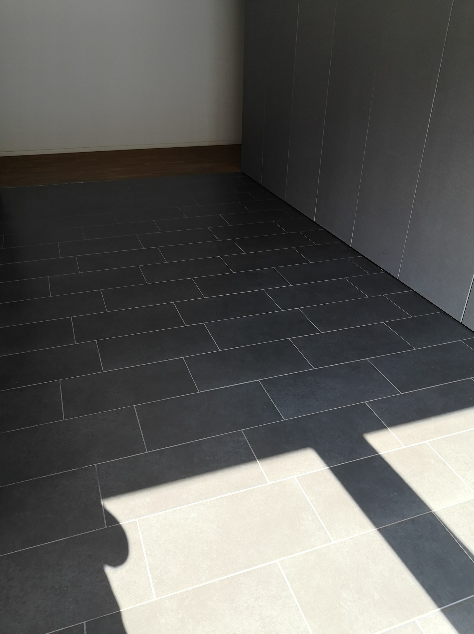 Ułożona grafitowa podłoga z płytek w nowym pomieszczeniu, widoczny fragment drewnianej podłogi w tle i cień padający na płytki.