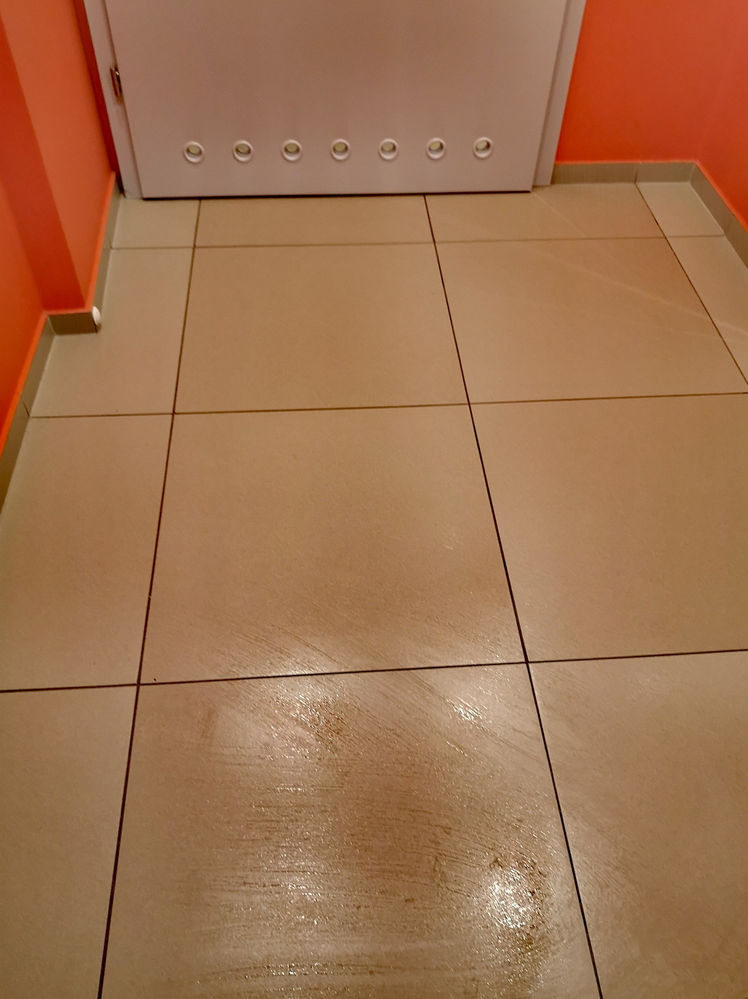 Beżowe płytki podłogowe po umyciu, widoczne mokre smugi, pomarańczowa ściana i białe drzwi z otworami wentylacyjnymi u góry.