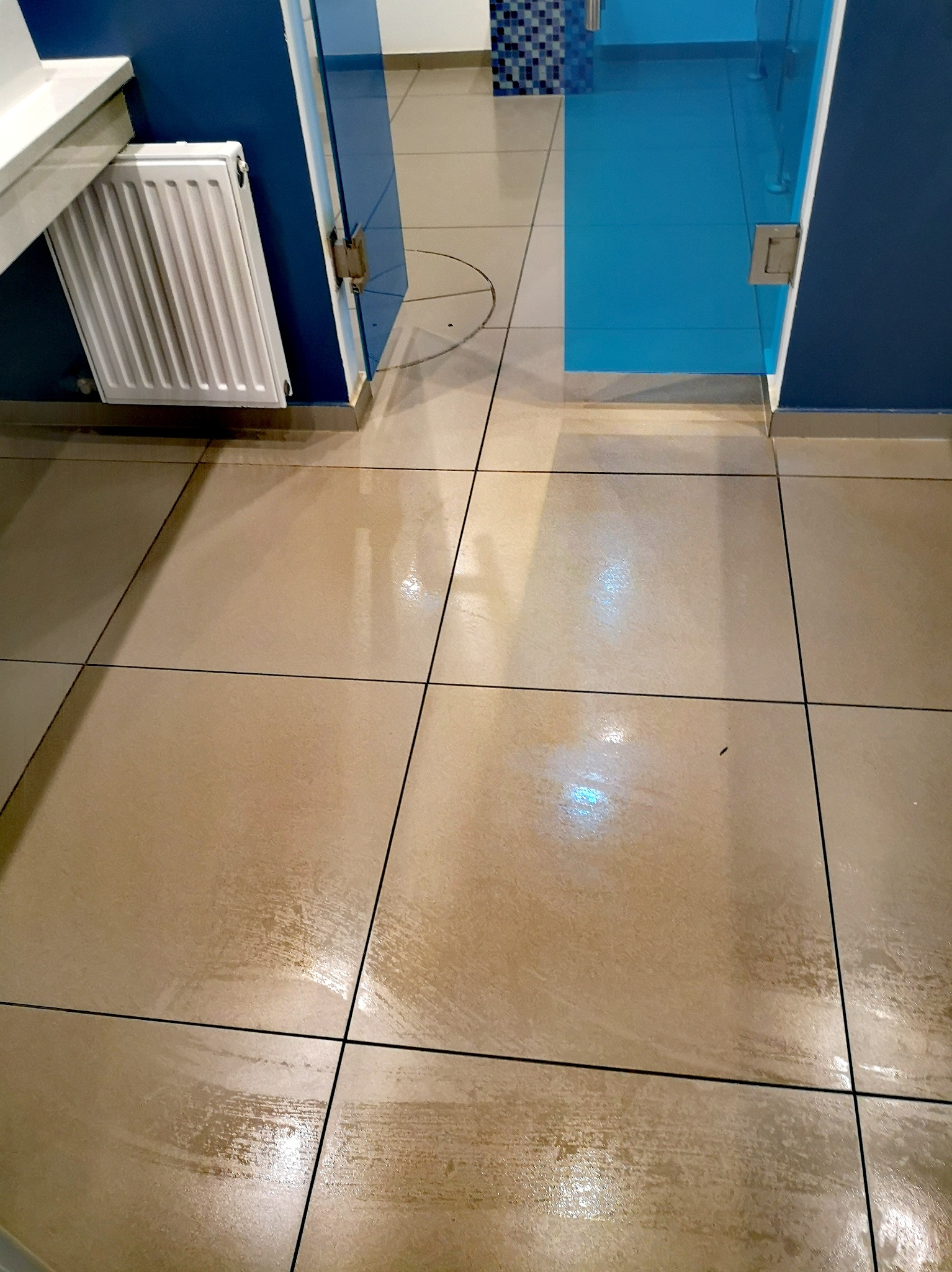 Błyszcząca, umyta podłoga z płytek w biurze, widoczny fragment grzejnika i przeszklone drzwi do pomieszczenia z niebieskimi ścianami.