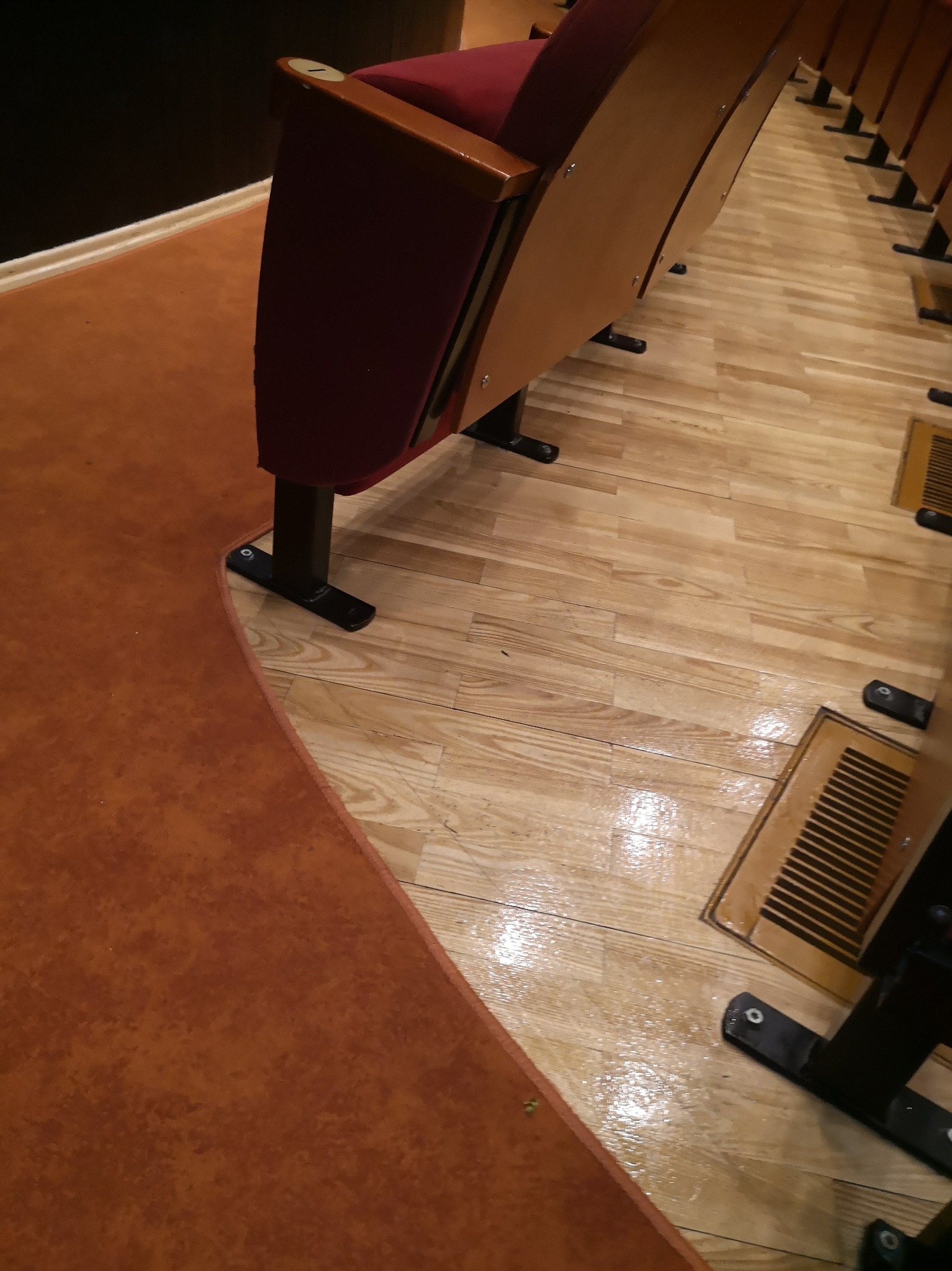Rząd foteli kinowych z bordową tapicerką, zamontowanych na drewnianej podłodze z kratkami wentylacyjnymi, przylegający do pomarańczowej wykładziny dywanowej