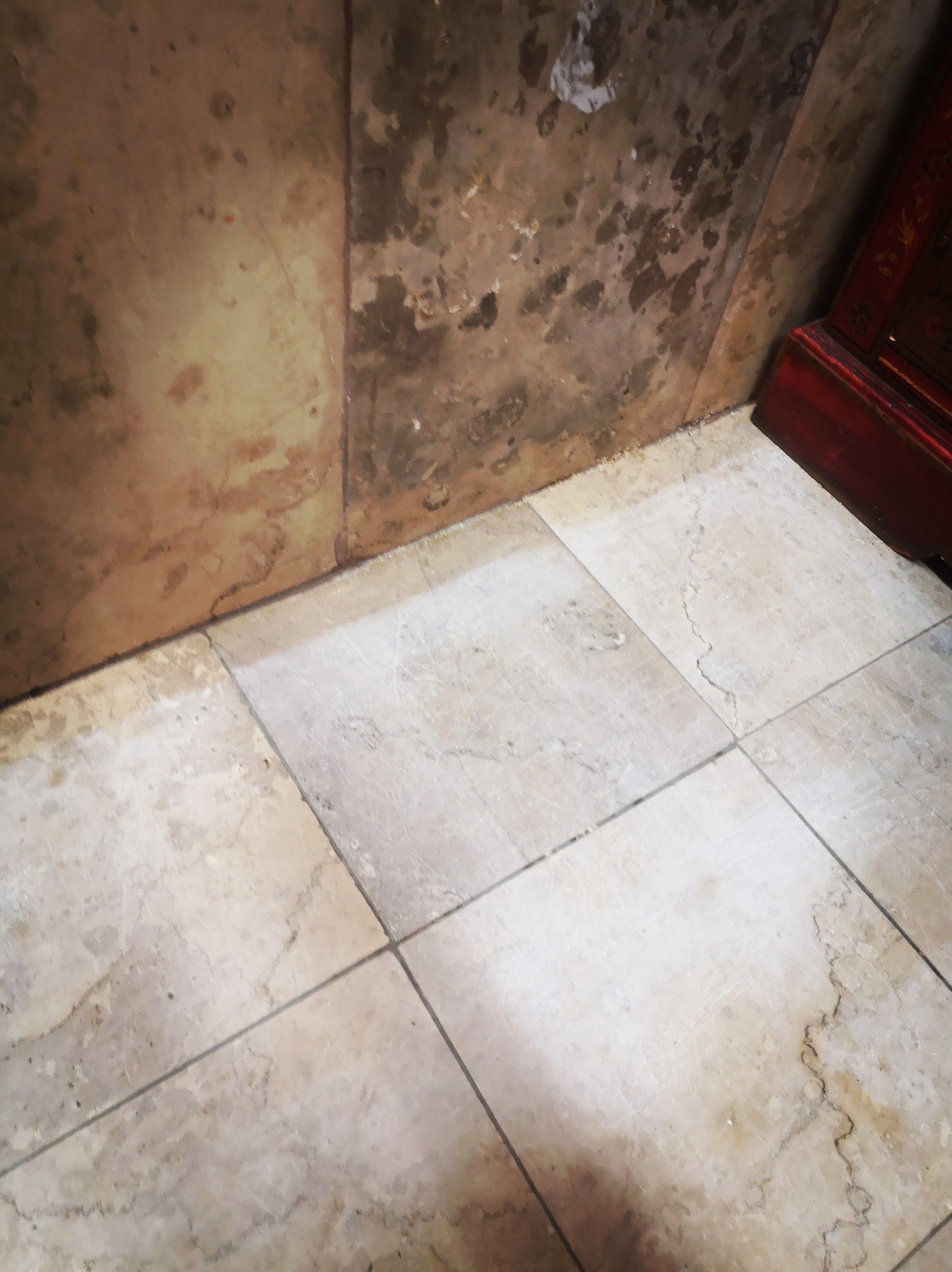 Brudna ściana z plamami i marmurowe płytki podłogowe wymagające czyszczenia, fragment czerwonego mebla w rogu.