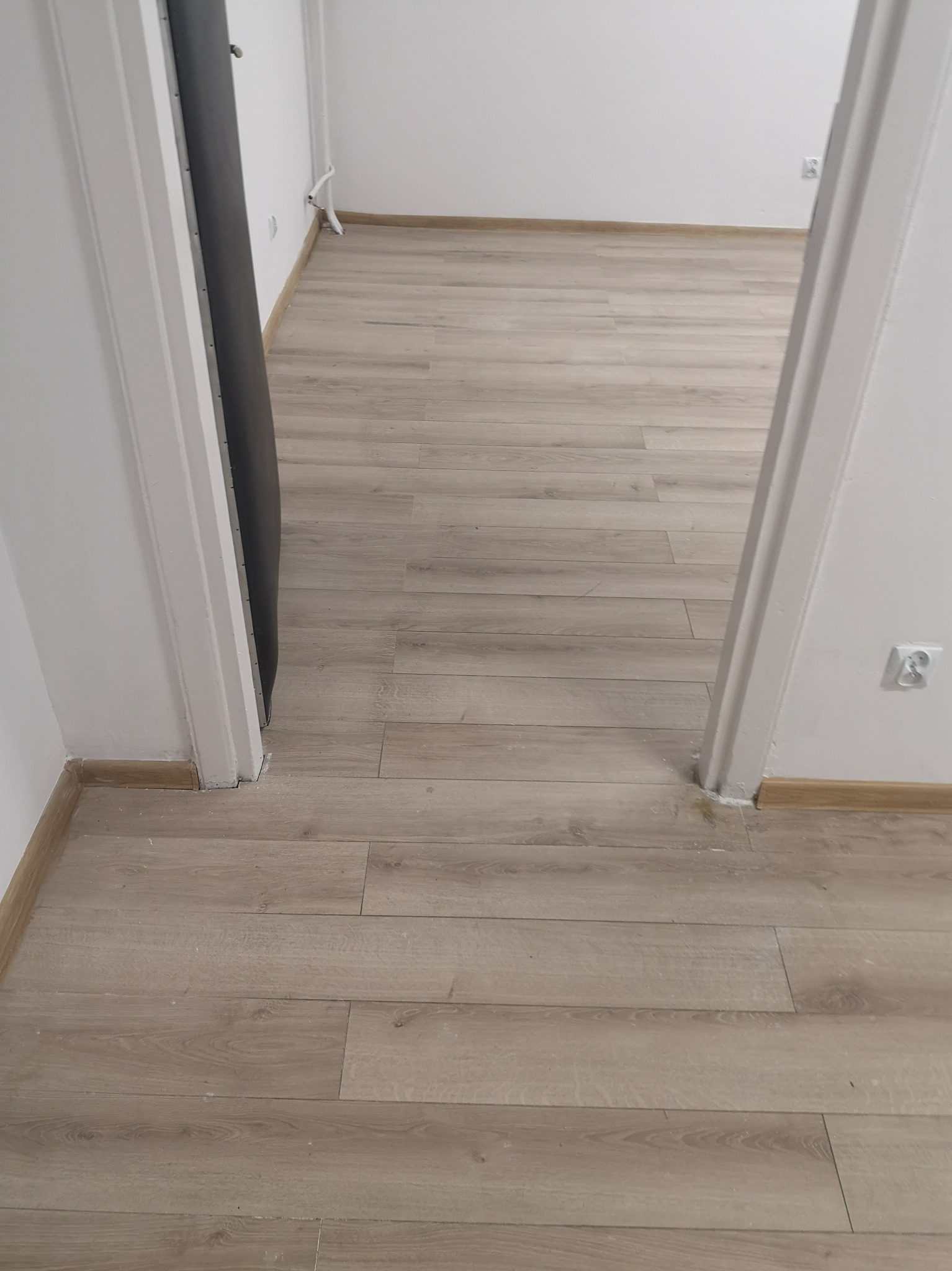 Ułożona podłoga z paneli w odcieniach jasnego drewna, widok z perspektywy wejścia, białe ściany i listwy przypodłogowe, widoczny fragment ościeżnicy drzwi.