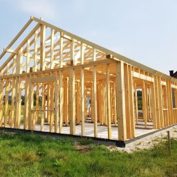 Krzysztof Rytel Domy - Surowy szkielet konstrukcji domu jednorodzinnego z drewna, widok z zewnątrz na tle trawnika i sąsiedniego budynku.