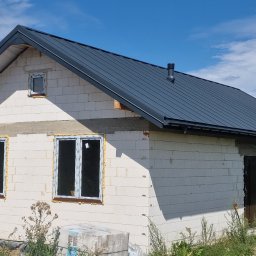 Krzysztof Rytel Domy - Niedokończony dom z białego betonu komórkowego z zamontowanymi oknami i grafitowym dachem z blachy na tle błękitnego nieba z chmurami.