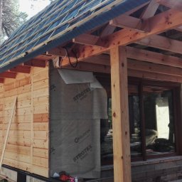 Krzysztof Rytel Domy - Budowa domu szkieletowego: drewniana konstrukcja ścian z deskowaniem, folia izolacyjna, widoczne elementy więźby dachowej i okna. Prace w toku, otoczenie leśne.