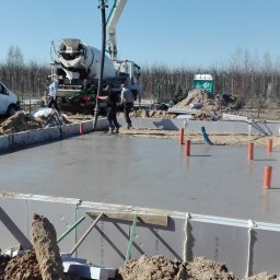 Krzysztof Rytel Domy - Wylewanie fundamentów pod budynek z betonomieszarki. Widoczne szalunki z izolacją termiczną, ekipa budowlana i częściowo zalany betonem teren budowy.