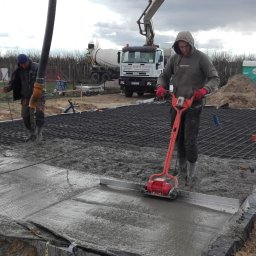 Krzysztof Rytel Domy - Prace betonowe: Dwóch robotników przy wylewaniu fundamentów pod budynek. Jeden używa wibratora do betonu, w tle betoniarka i rozległy teren budowy.