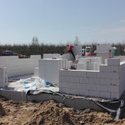 Krzysztof Rytel Domy - Wznoszenie ścian parteru z białych bloczków na fundamencie. Widoczny pracownik w czerwonej koszuli, częściowo zbudowane ściany i teren budowy z piaskiem.