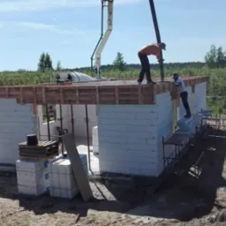 Budowa domu z betonu komórkowego: dwóch robotników w trakcie wylewania stropu przy użyciu pompy do betonu. Widok na częściowo wzniesione ściany i zielone otoczenie.
