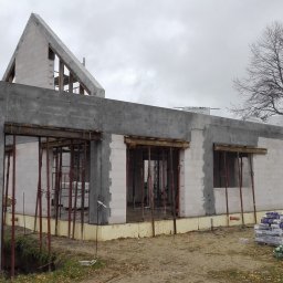 Krzysztof Rytel Domy - Budowa domu w stanie surowym otwartym: ściany z betonu komórkowego, betonowe słupy, widoczne zbrojenia okien, materiały budowlane na ziemi, pochmurne niebo.