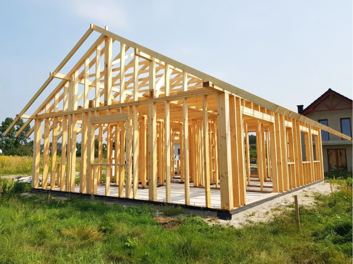 Surowy szkielet konstrukcji domu jednorodzinnego z drewna, widok z zewnątrz na tle trawnika i sąsiedniego budynku.