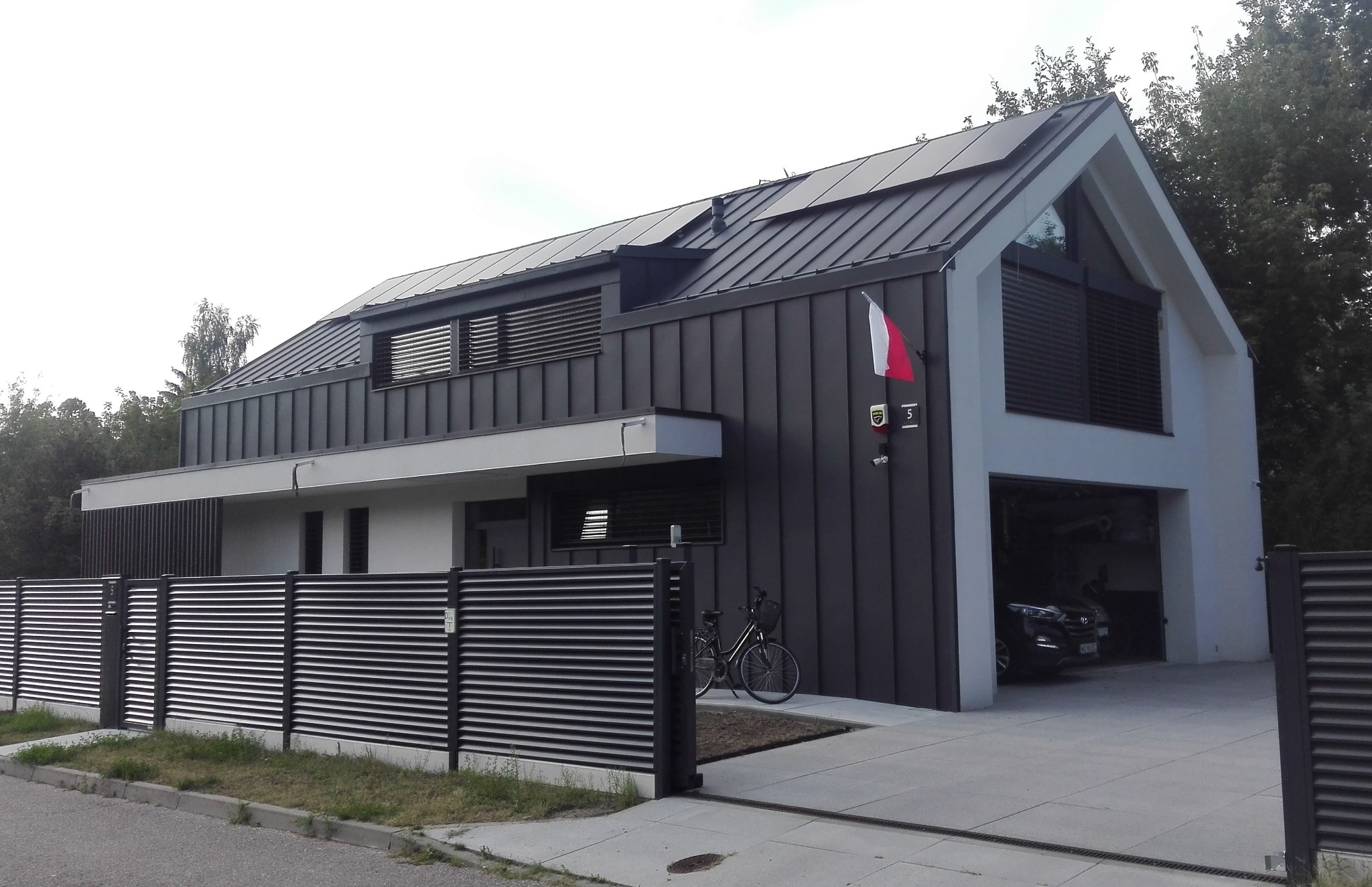 Nowoczesny dom z ciemną, metalową elewacją, ukośnym dachem z panelami słonecznymi, garażem w bryle budynku i metalowym ogrodzeniem lamelowym.