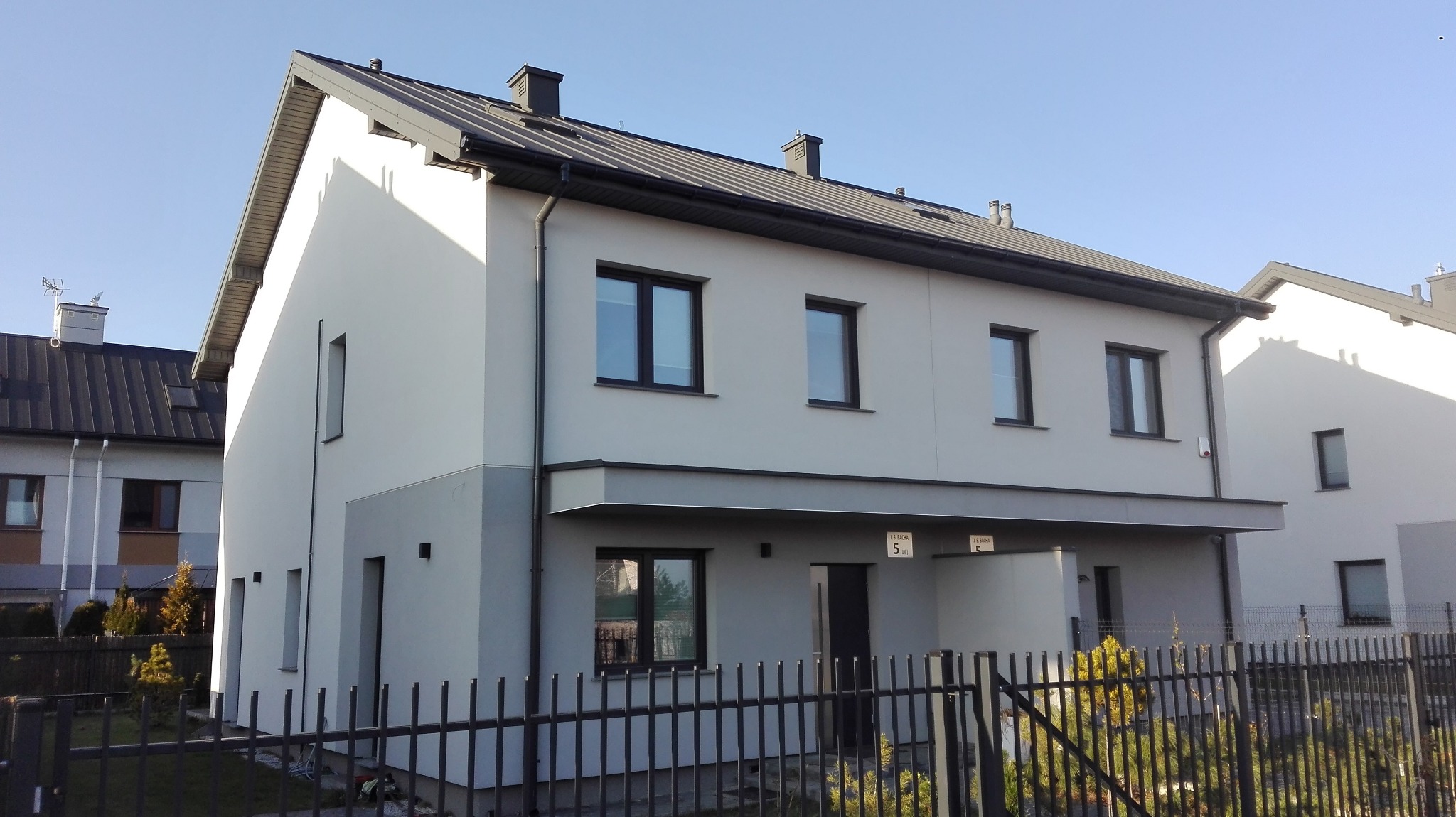 Dwukondygnacyjny budynek mieszkalny w odcieniach szarości z czarną stolarką okienną i dachem z blachy, widziany zza metalowego ogrodzenia.