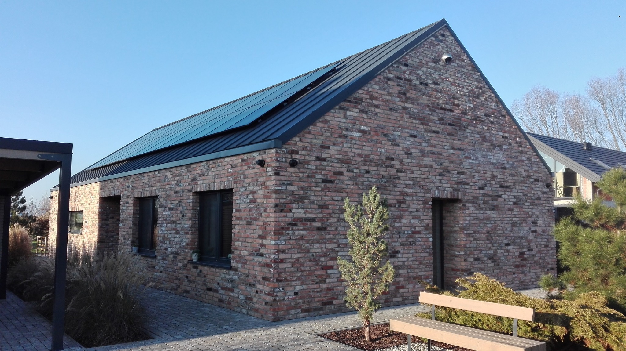 Jednopiętrowy dom z czerwonej cegły klinkierowej z panelami słonecznymi na dachu, małe drzewko iglaste przed budynkiem, ławka i kostka brukowa na pierwszym planie.