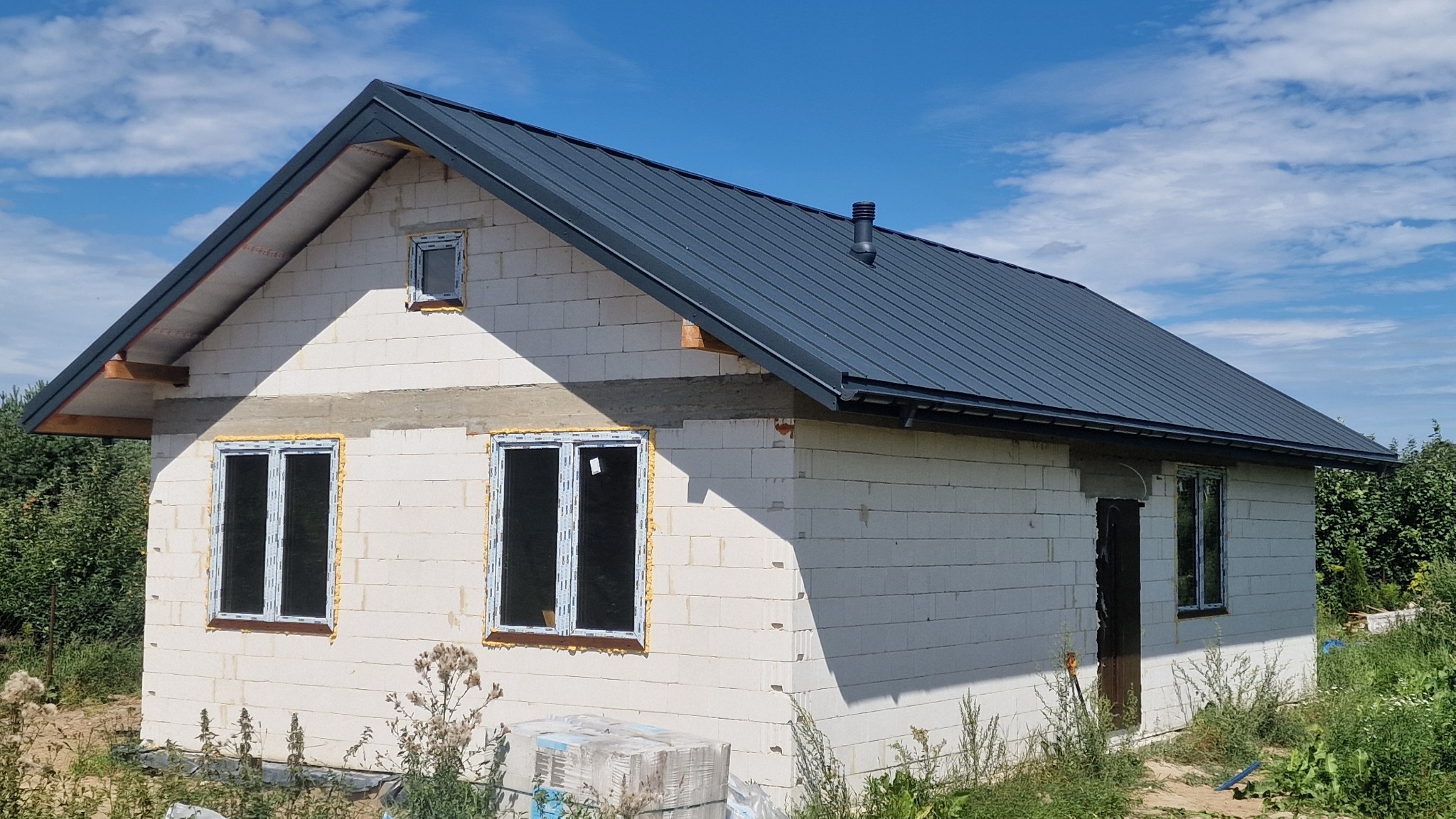 Niedokończony dom z białego betonu komórkowego z zamontowanymi oknami i grafitowym dachem z blachy na tle błękitnego nieba z chmurami.