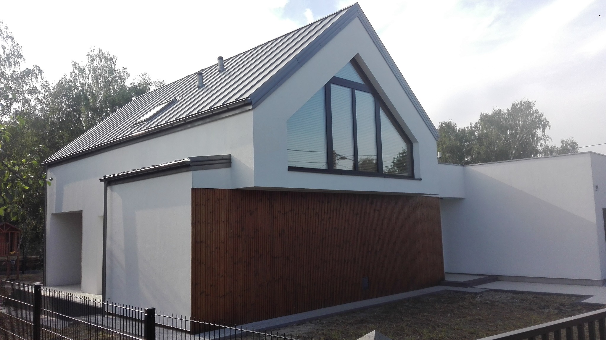 Nowoczesny dom z drewnianą elewacją i trójkątnym oknem na poddaszu, otoczony zielenią. Metalowy dach z widocznymi oknami połaciowymi.