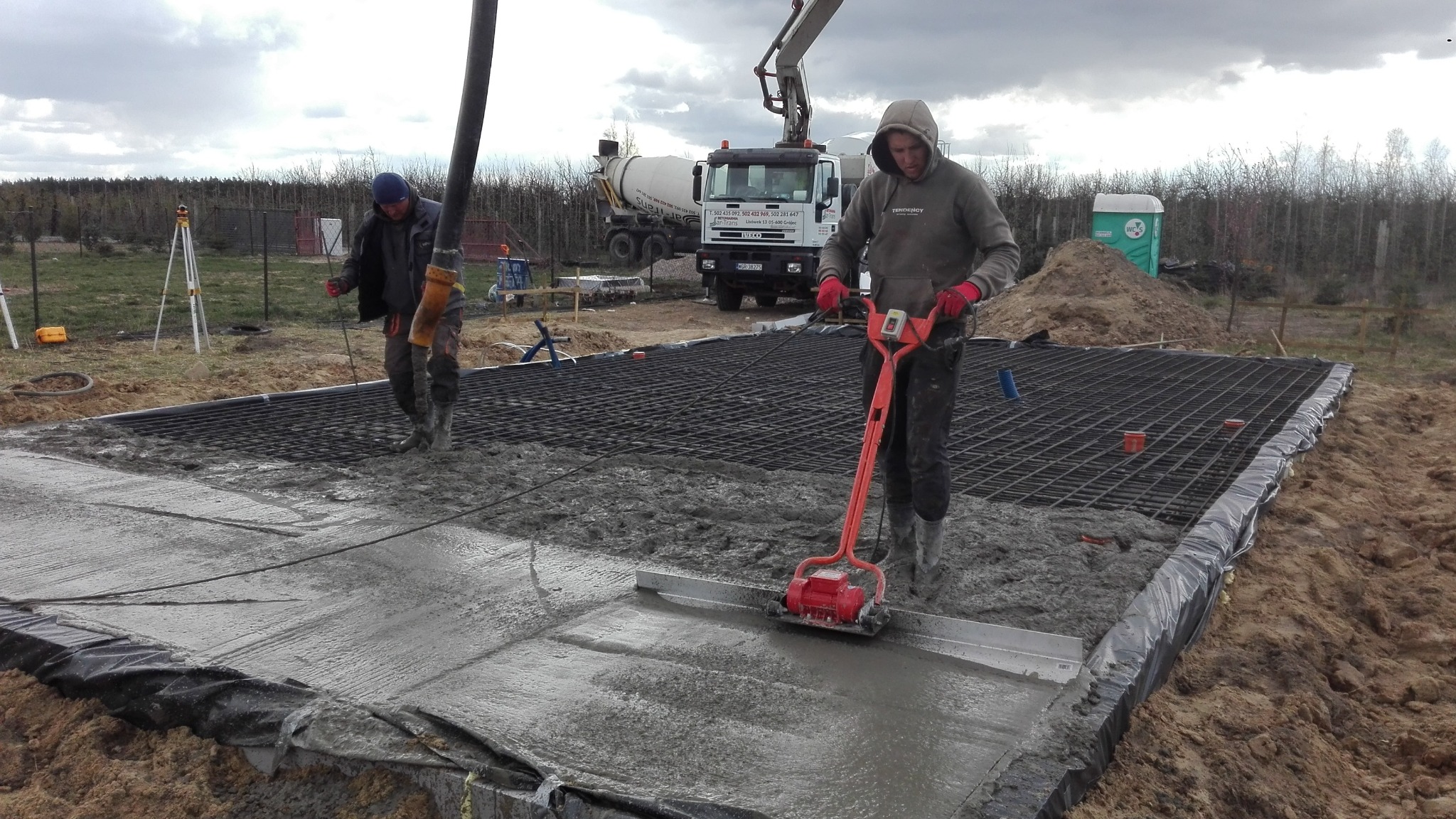 Prace betonowe: Dwóch robotników przy wylewaniu fundamentów pod budynek. Jeden używa wibratora do betonu, w tle betoniarka i rozległy teren budowy.