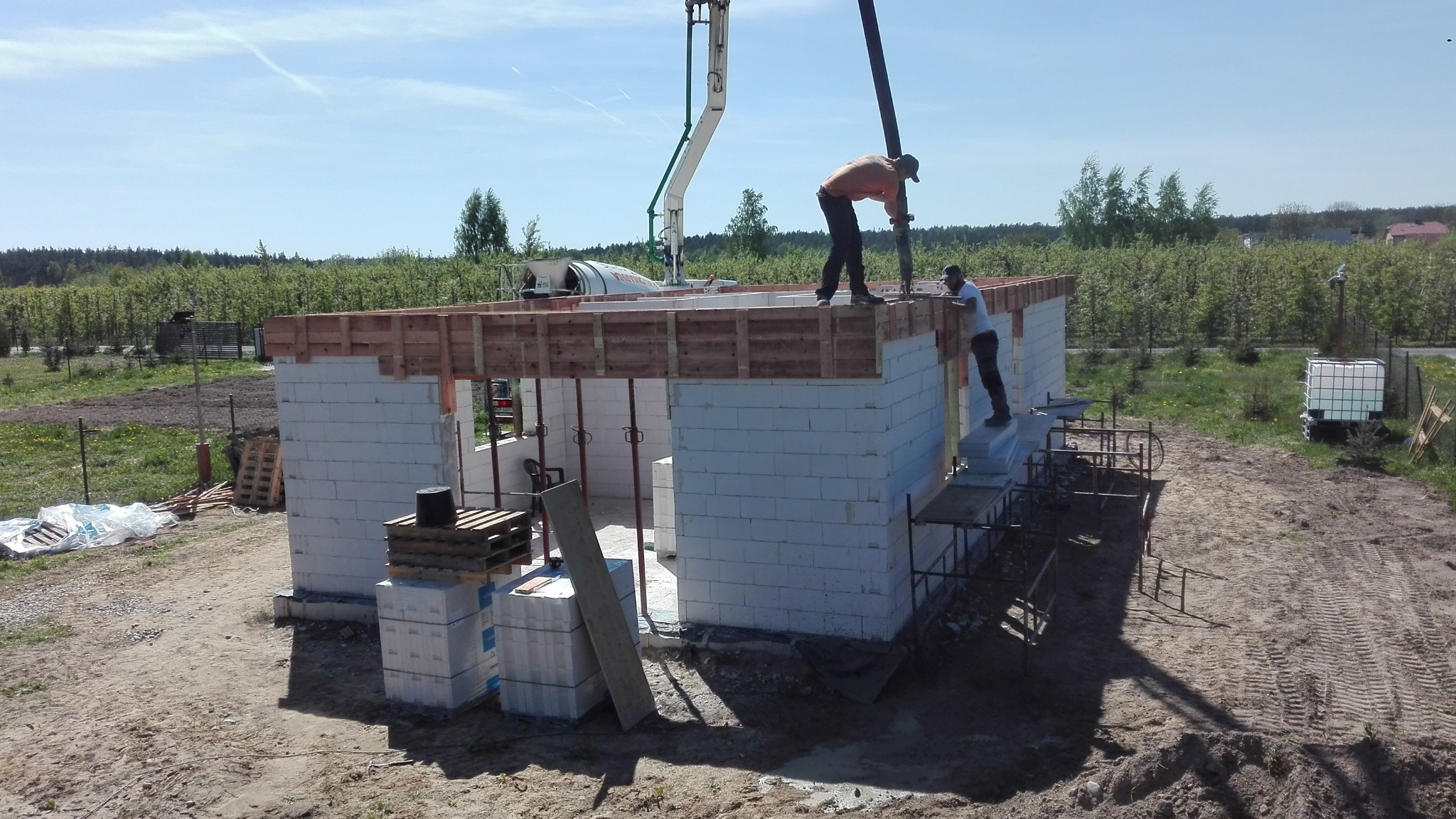 Budowa domu z betonu komórkowego: dwóch robotników w trakcie wylewania stropu przy użyciu pompy do betonu. Widok na częściowo wzniesione ściany i zielone otoczenie.