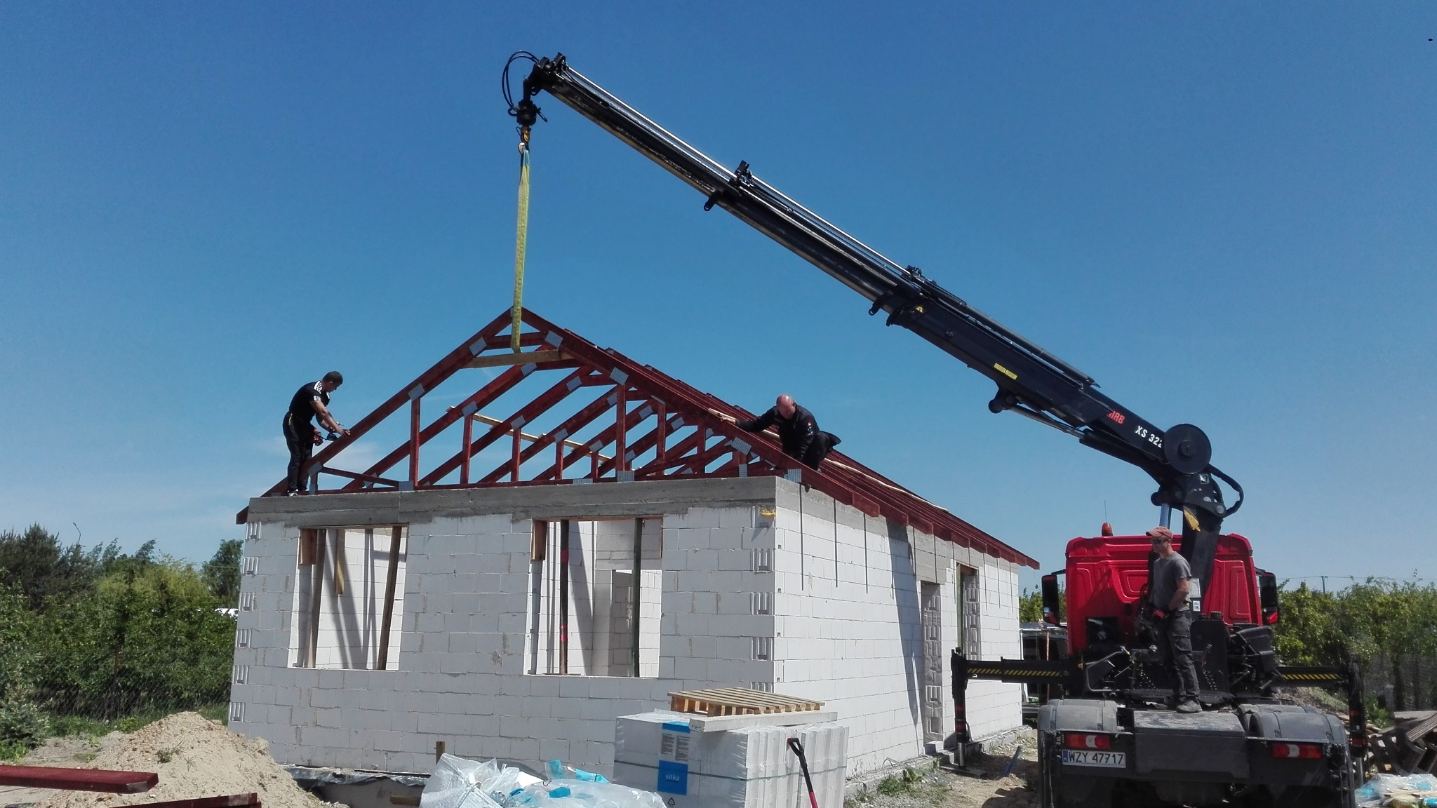 Wznoszenie konstrukcji dachu domu z białych bloczków. Żuraw podnosi element więźby dachowej, a robotnicy montują go na szczycie budynku w słoneczny dzień.