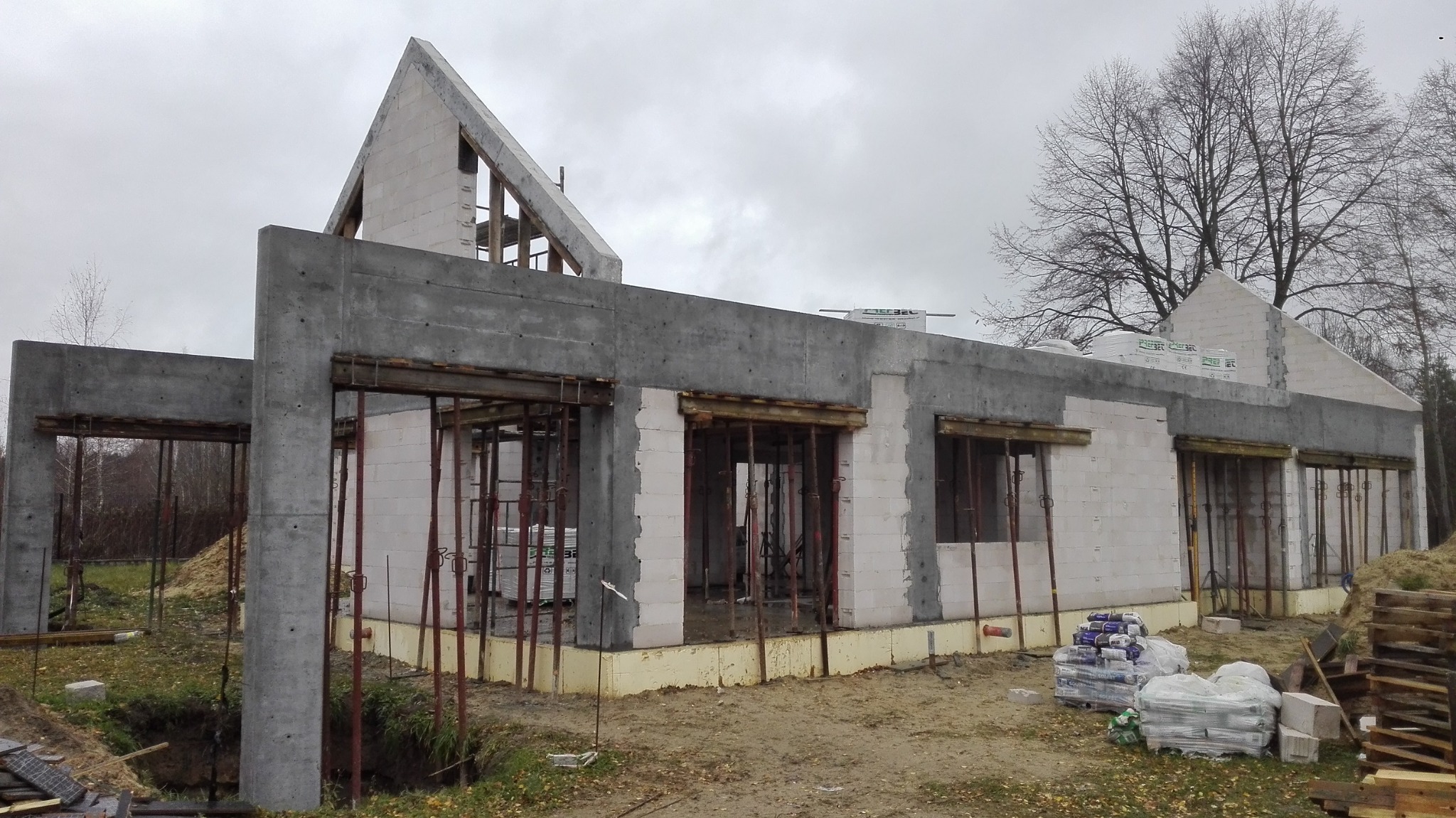 Budowa domu w stanie surowym otwartym: ściany z betonu komórkowego, betonowe słupy, widoczne zbrojenia okien, materiały budowlane na ziemi, pochmurne niebo.