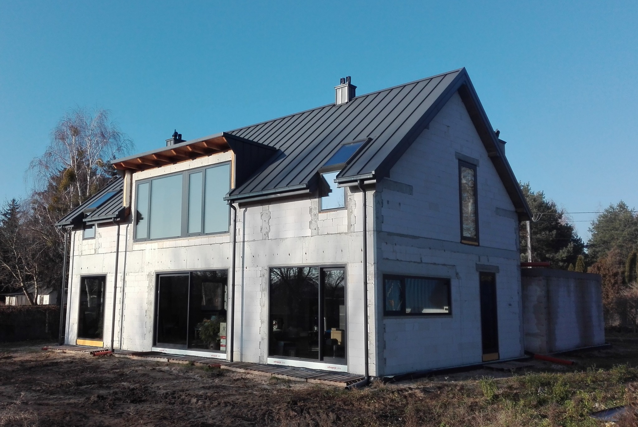 Niedokończony dom z białego bloczka z dużymi oknami i ciemnym dachem na tle błękitnego nieba. Widoczne elementy konstrukcyjne i teren budowy.