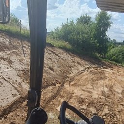 Kamil Warkocki Usługi Ziemne - Widok z kabiny maszyny budowlanej na świeżo wyrównany, wilgotny teren pod budowę, z widocznymi śladami opon i roślinnością w tle.