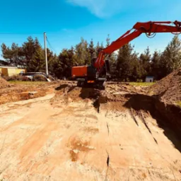 Czerwona koparka na placu budowy, przygotowany wykop pod fundamenty, otoczenie leśne, błękitne niebo.