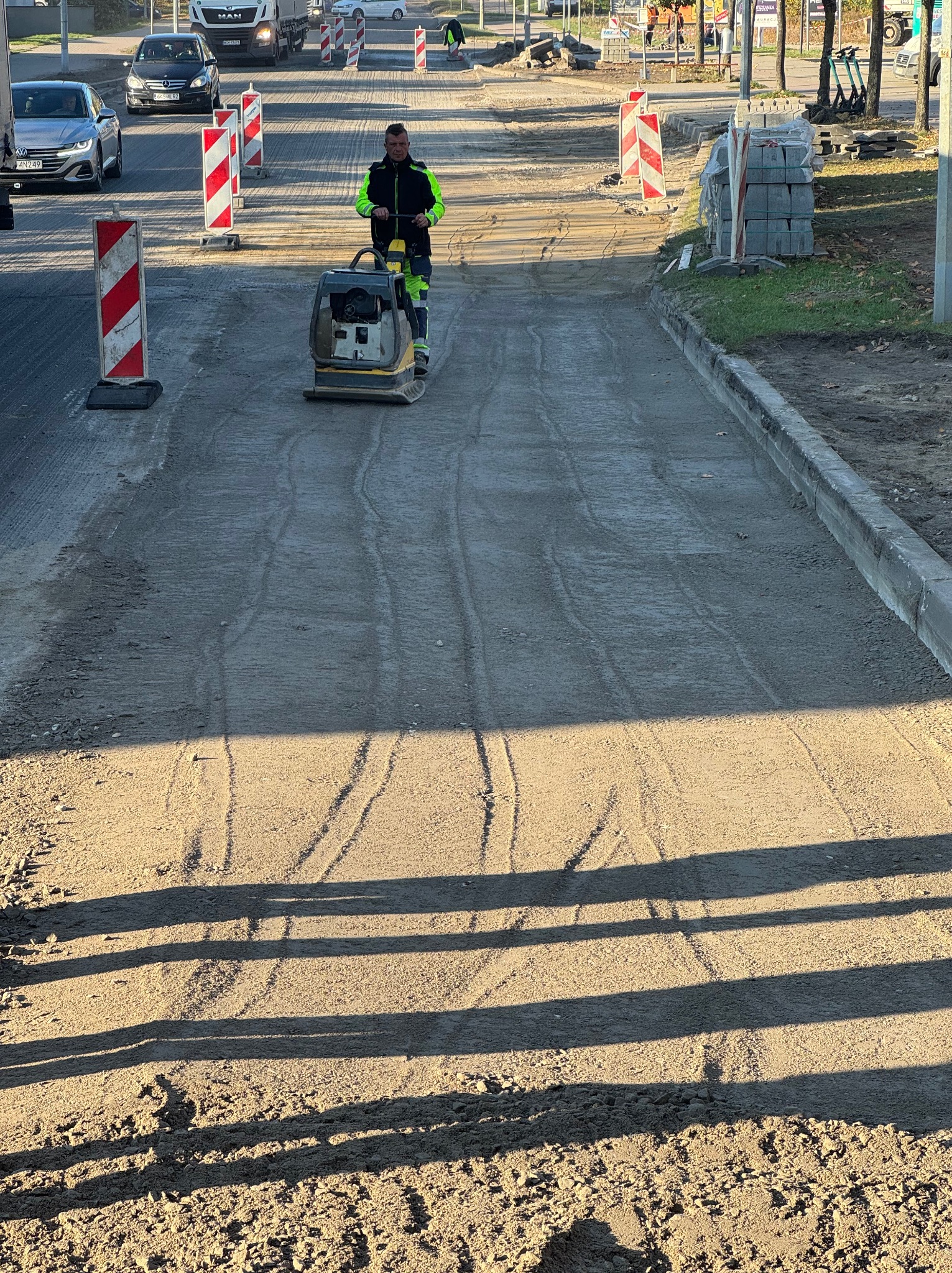 Pracownik w odblaskowej kurtce zagęszcza podłoże pod chodnik w trakcie budowy drogi, widoczne ustawione bariery drogowe i palety z kostką brukową