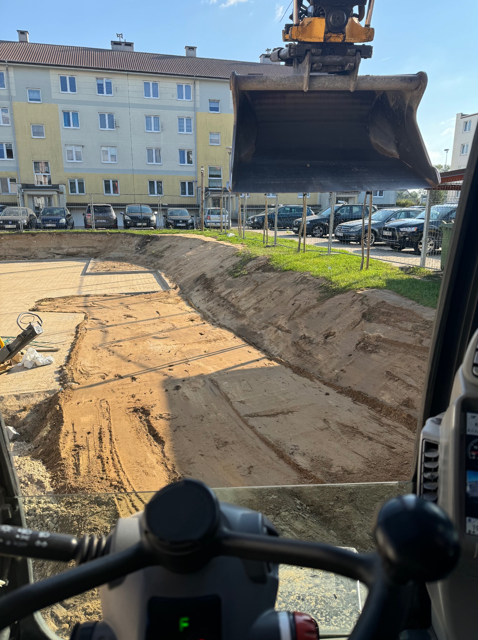 Widok z kabiny koparki na wykop pod budynek, zarys fundamentów widoczny w piasku, w tle budynek mieszkalny i zaparkowane samochody.