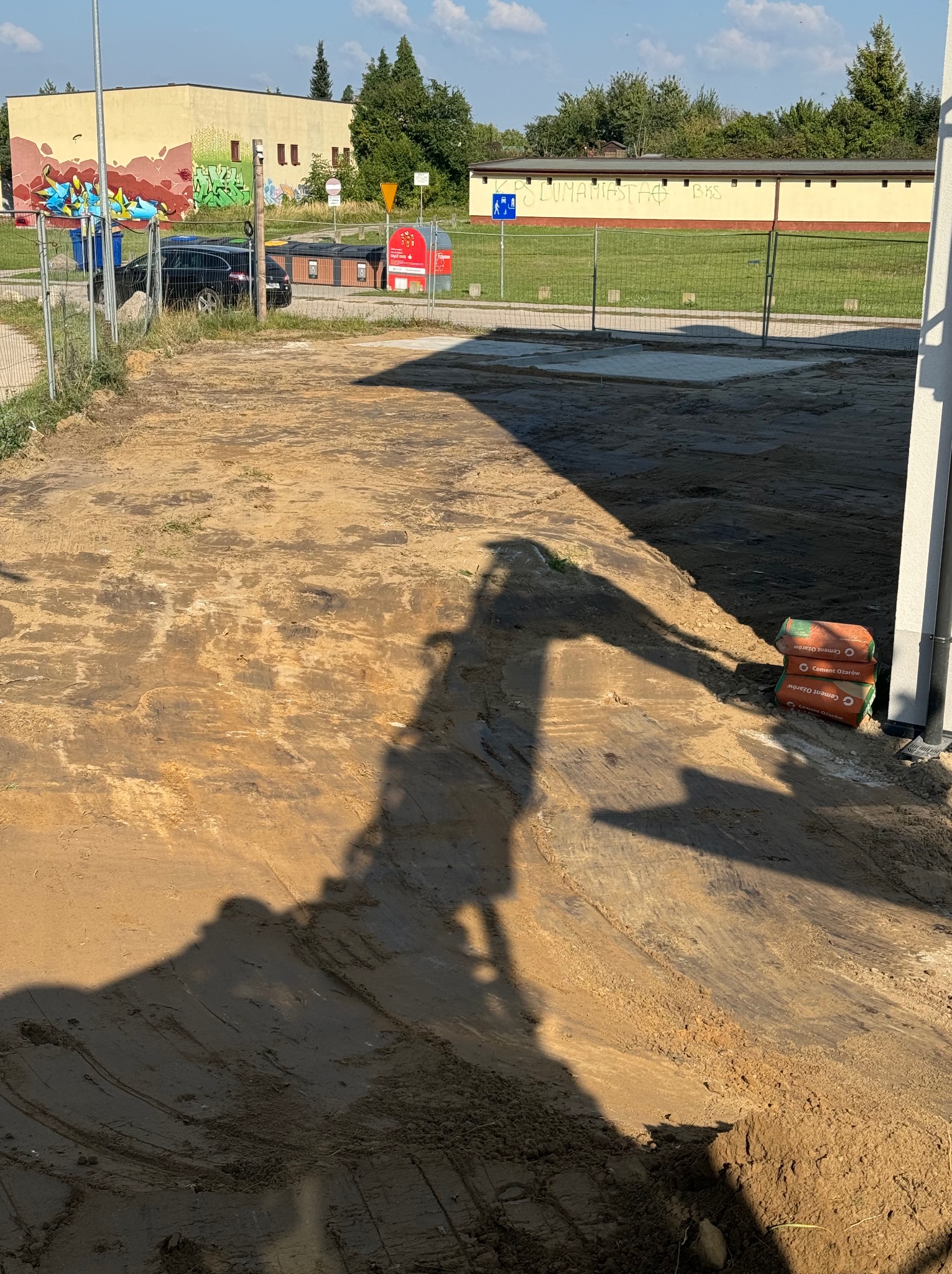 Przygotowany teren pod budowę, widoczny cień koparki, obok paleta z workami cementu, w tle budynek z graffiti.