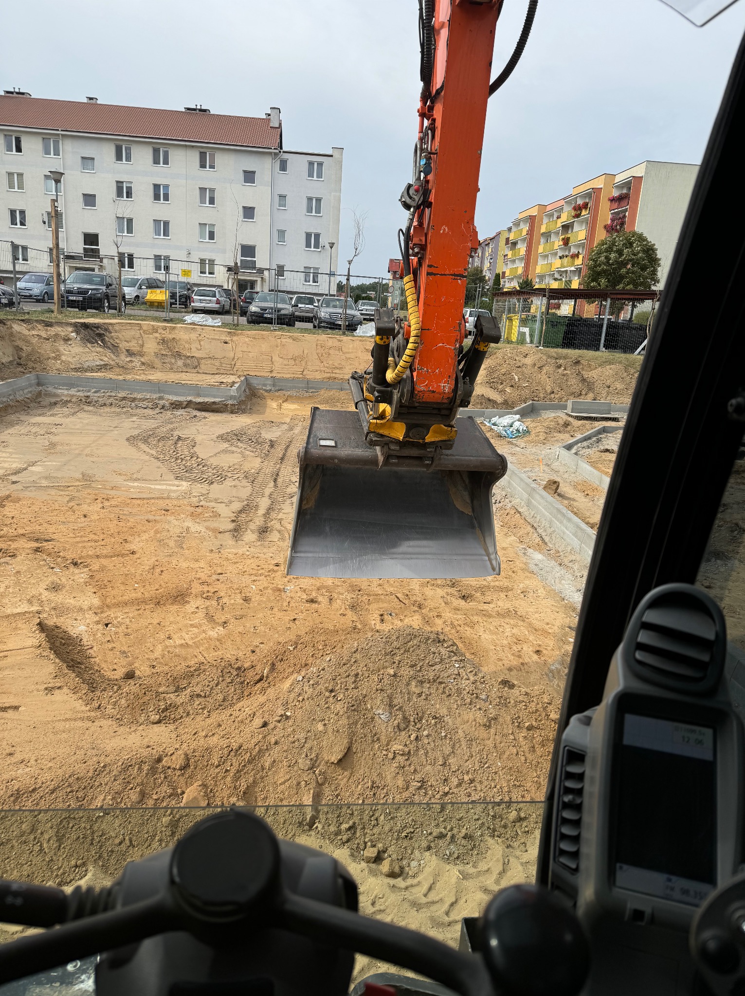 Widok z kabiny koparki na wykop pod fundamenty budynku mieszkalnego, z widocznymi betonowymi ławami fundamentowymi i hałdą piasku.