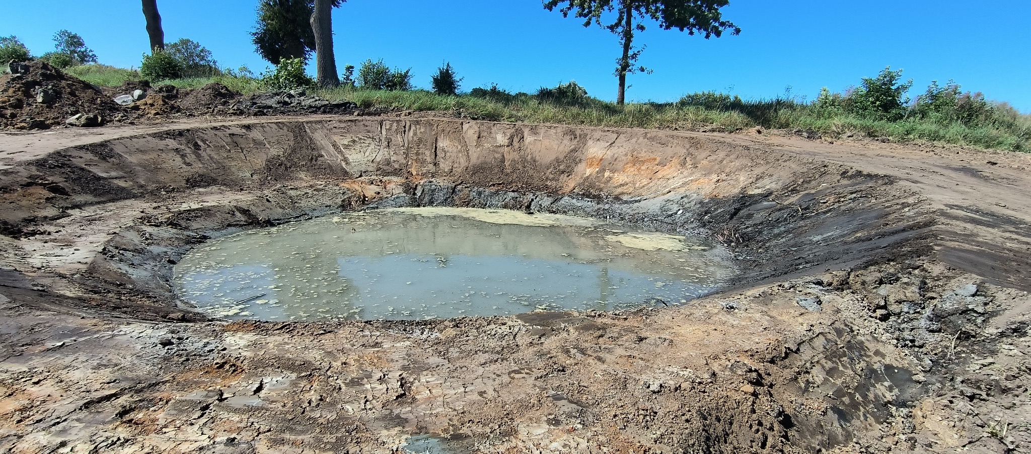 Okrągły wykop ziemny wypełniony wodą z zielonkawym nalotem, otoczony ubitą ziemią i zielenią w tle, wykonany w słoneczny dzień.