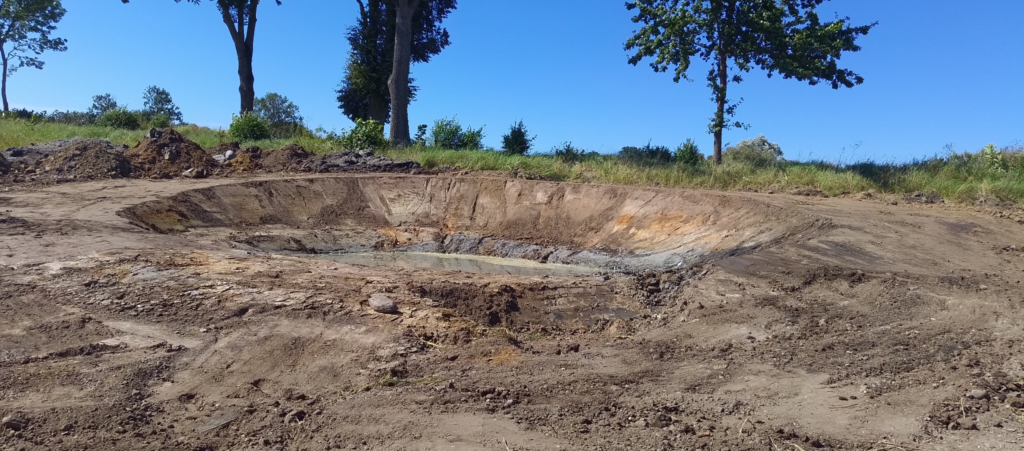 Wykopany dół pod staw z wodą na dnie, otoczony ziemią i trawą, widoczne drzewa w tle pod błękitnym niebem.