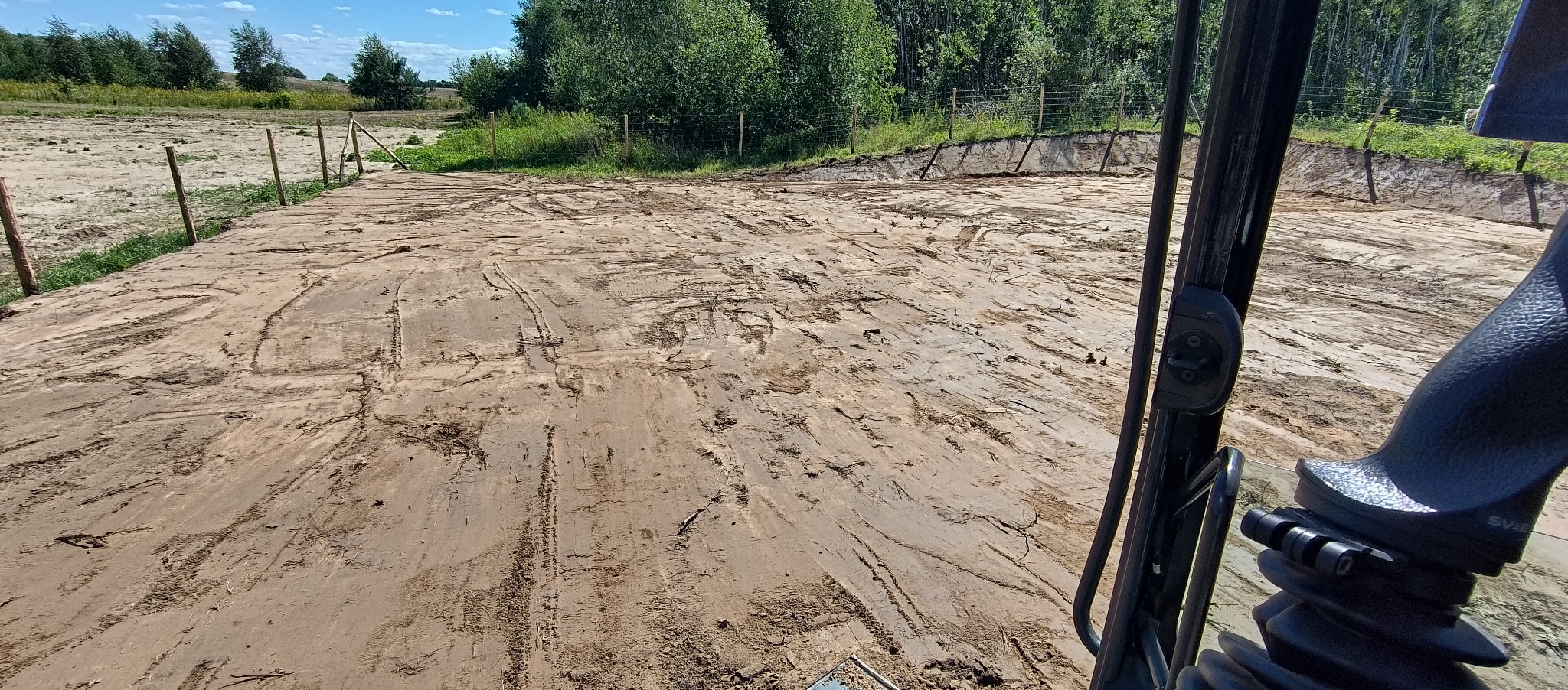 Widok z kabiny na wyrównany teren piaszczysty z widocznymi śladami opon, ogrodzony drewnianymi słupkami, z zielenią w tle.