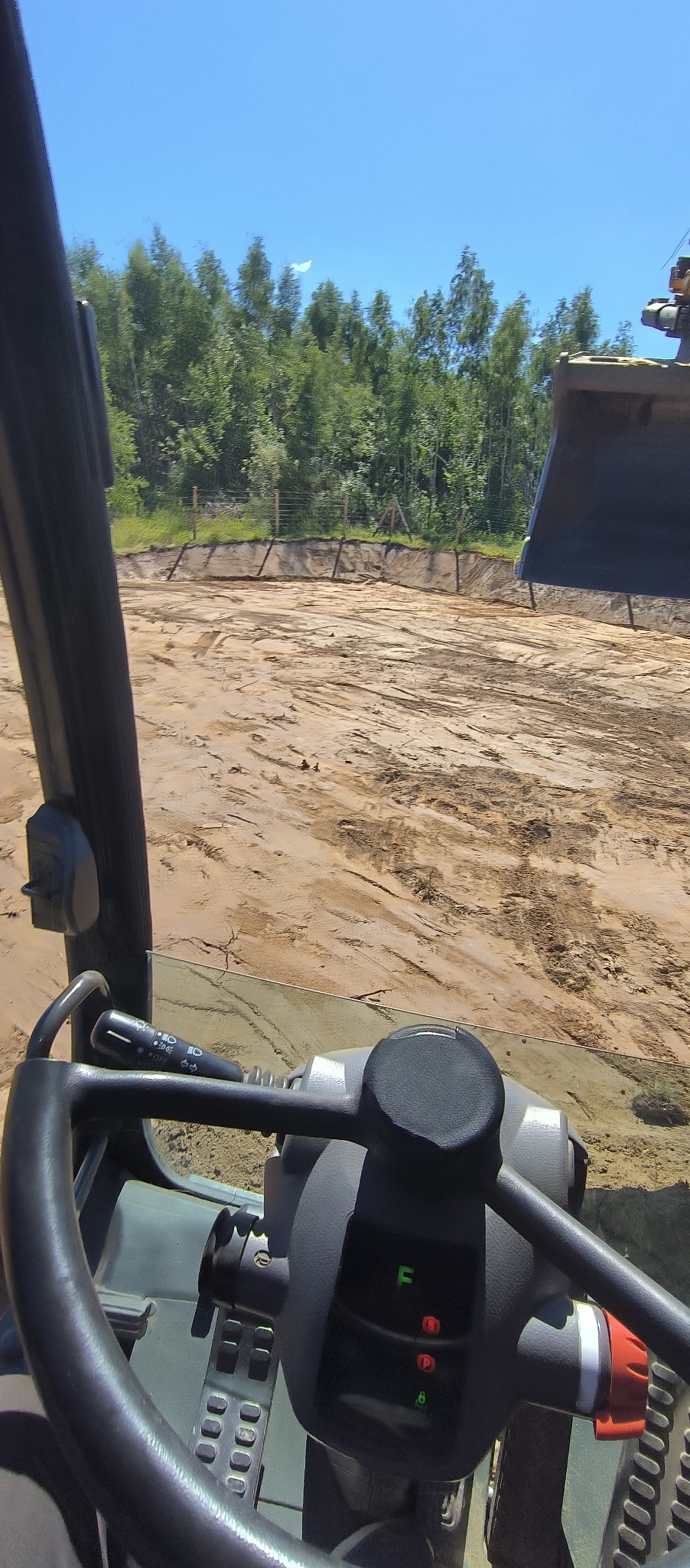 Widok z kabiny maszyny budowlanej na wyrównany teren, w tle zielone drzewa i błękitne niebo.