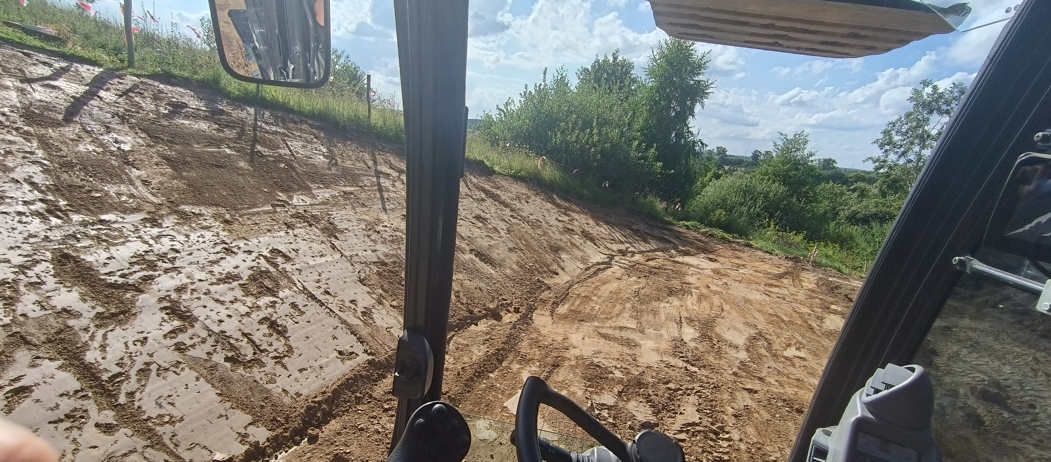 Widok z kabiny maszyny budowlanej na świeżo wyrównany, wilgotny teren pod budowę, z widocznymi śladami opon i roślinnością w tle.