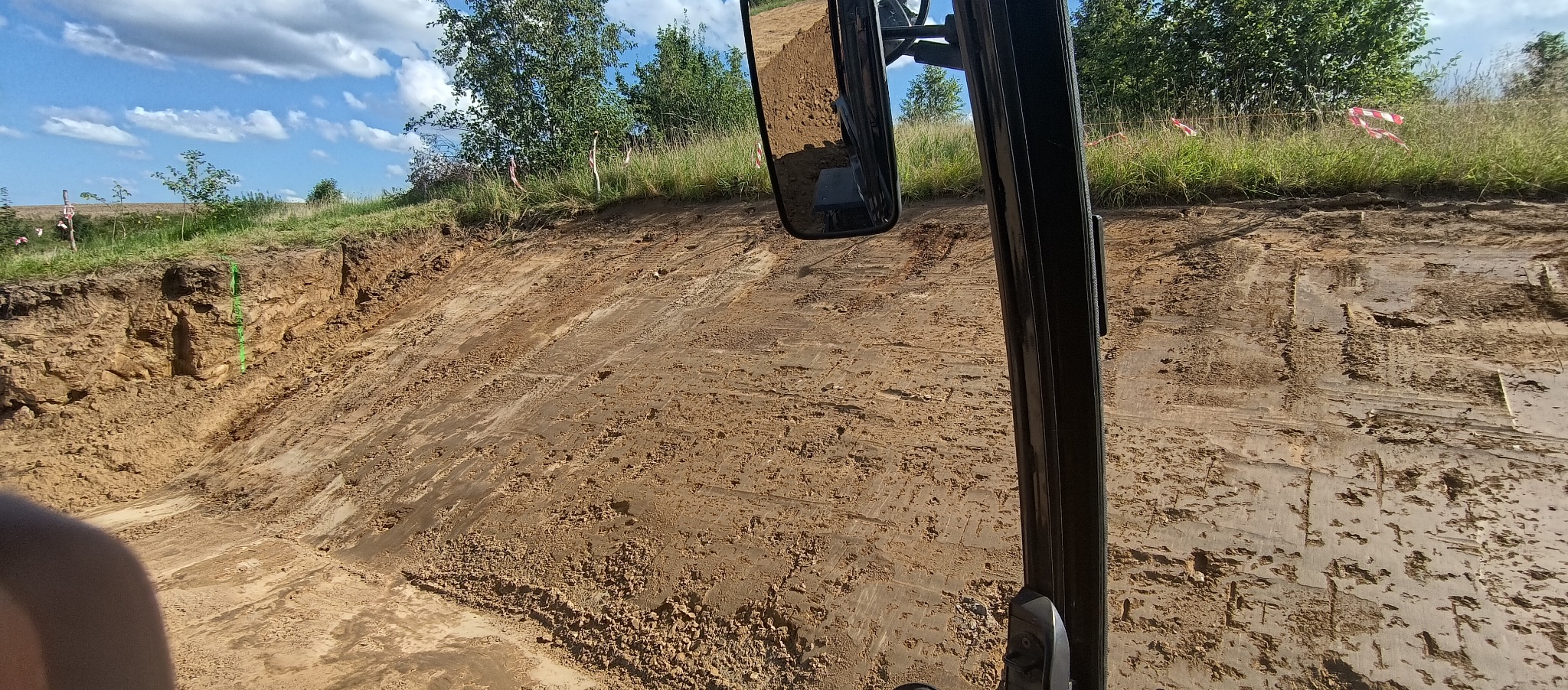 Wyrobisko w ziemi z widocznymi śladami opon, częściowo zarośnięte trawą, z perspektywy kabiny maszyny budowlanej, widoczne niebo z chmurami.