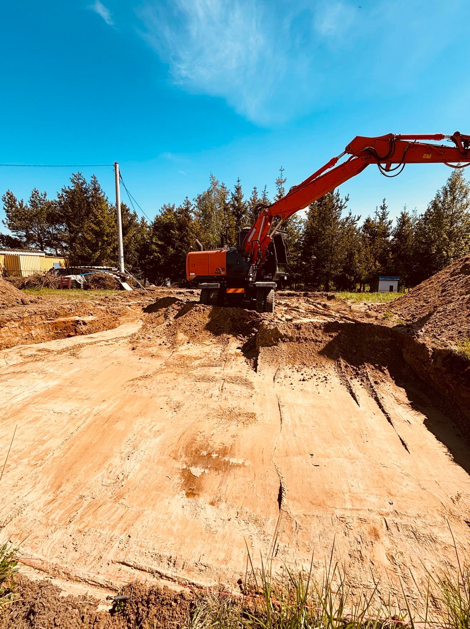 Czerwona koparka na placu budowy, przygotowany wykop pod fundamenty, otoczenie leśne, błękitne niebo.