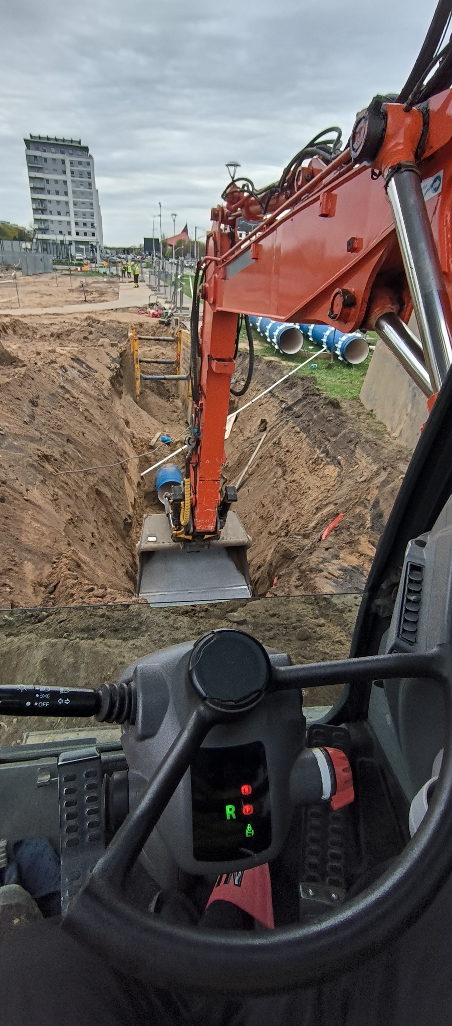 Widok z kabiny koparki na wykop pod fundamenty z zabezpieczeniami ścian wykopu, w tle budynek mieszkalny i rury kanalizacyjne.
