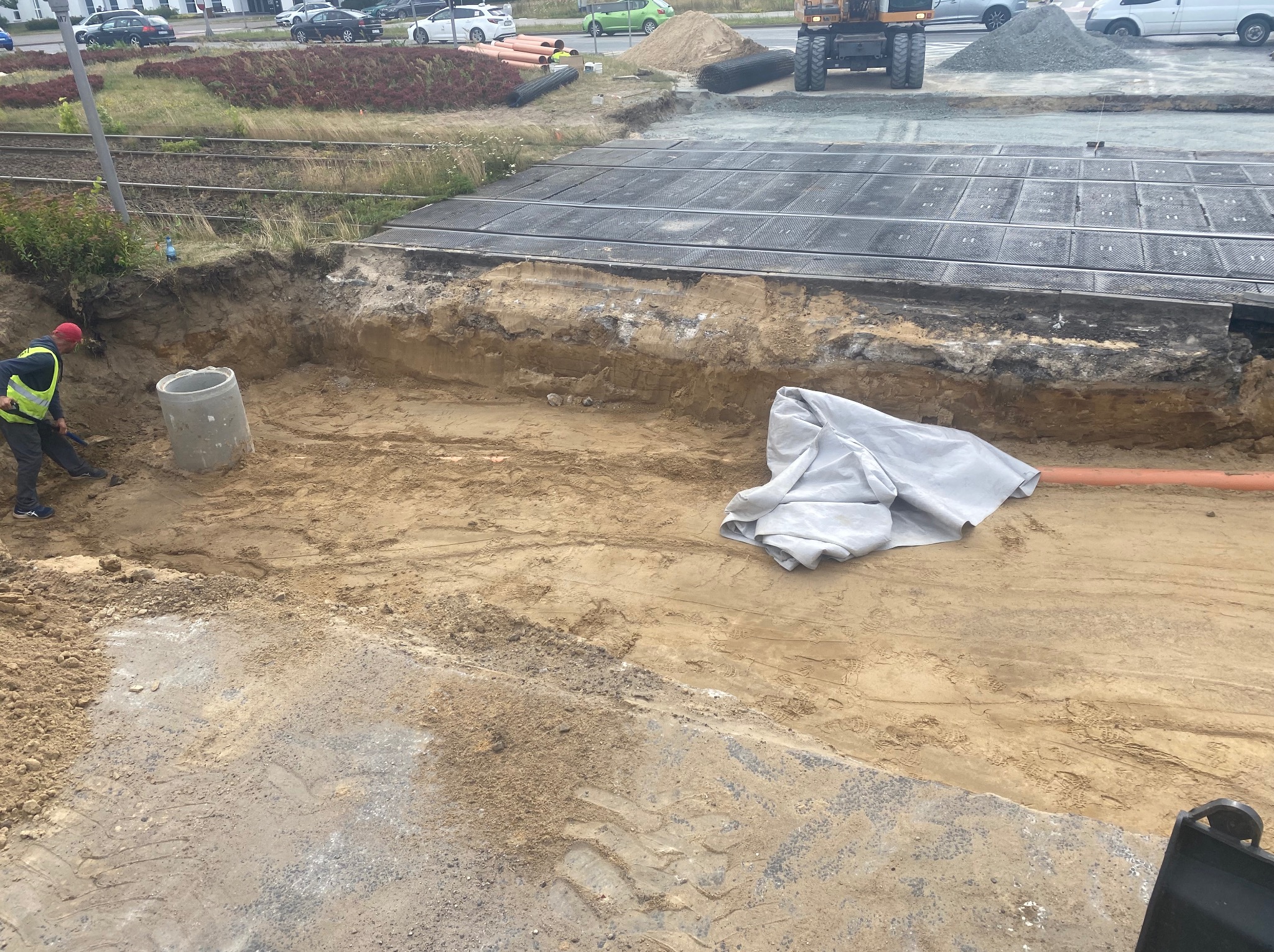 Wykop ziemny przy torach kolejowych z widocznym betonowym kręgiem, pomarańczową rurą i pracownikiem w odblaskowej kamizelce. W tle maszyna budowlana i materiały budowlane.